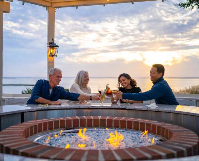 Four people gather around a circular fire pit, toasting drinks as the sun sets over a serene waterfront. Lanterns illuminate the scene.