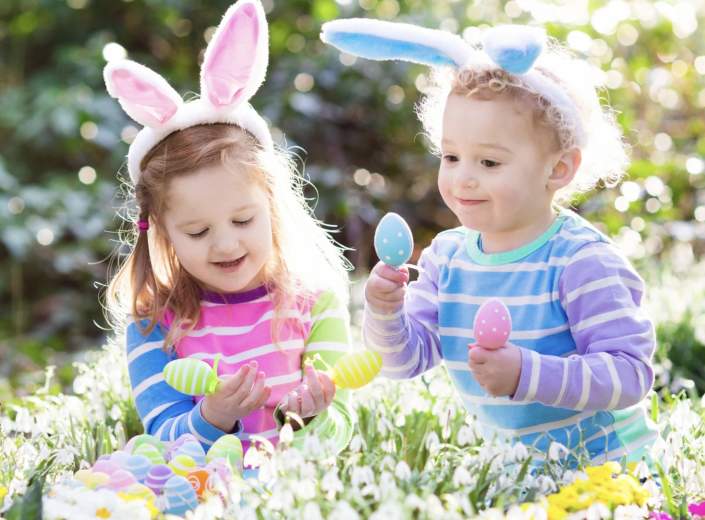Two young children in striped shirts, wearing bunny ear headbands, joyfully play with colourful Easter eggs in a sunny, flower-filled garden.