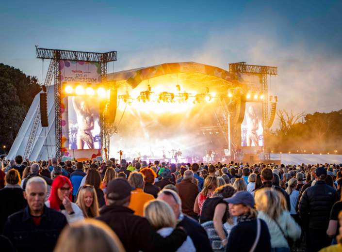 A large crowd of people enjoying an outdoor concert, with colourful lights and a stage in the background.