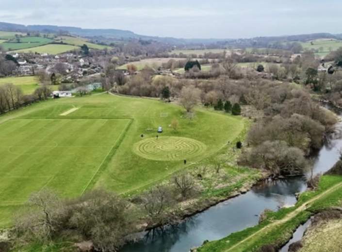 Aerial view of a countryside landscape featuring a sports field with goalposts, a circular grassy labyrinth, and a winding river. Rolling hills and scattered trees convey a peaceful, rural vibe.