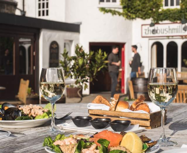 Plates of food and glasses of wine in the foreground of the image with two people in the background chatting, outside the front of The Bushmills Inn.