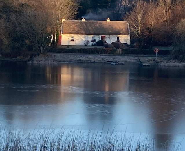 Keenaghan Cottage, a white thatched cottage, reflected in a calm, icy lake and surrounded by winter trees in soft morning light.