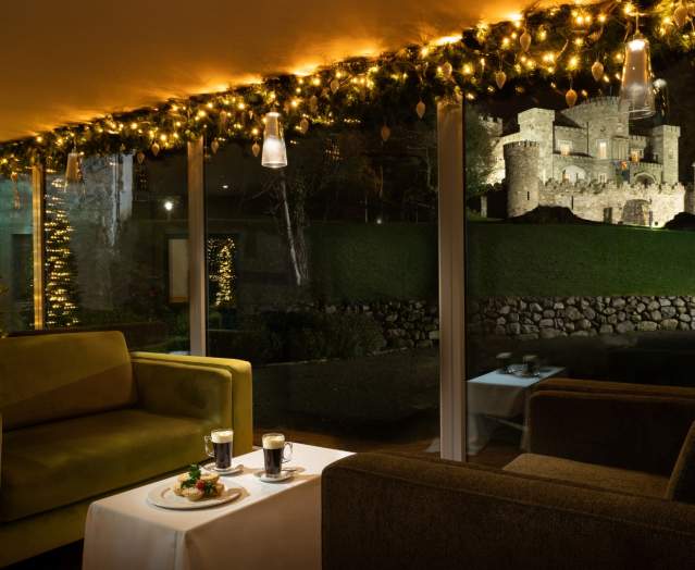 Cosy, festive lounge with warm lights and armchairs, looking out through large windows at the illuminated Killeavy Castle at night.