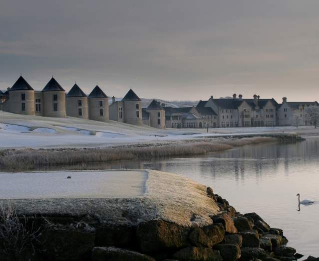 Snow-covered Lough Erne Resort reflected in a calm lake at winter sunrise, with two swans gliding across the water.