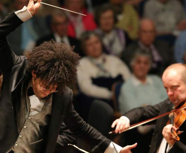 A musical conductor deep in conducting and a musician playing a violin with an audience behind them.