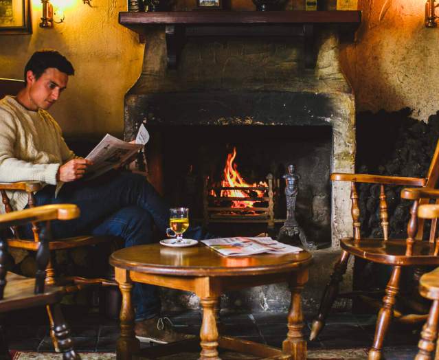 Man sits by the fire reading a newspaper with a hot whiskey on the table