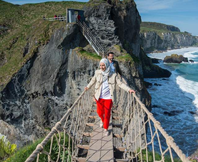 Stunning views of Carrick-a-Rede Rope Bridge, along the Causeway Coastal Route as a couple cross the bridge.