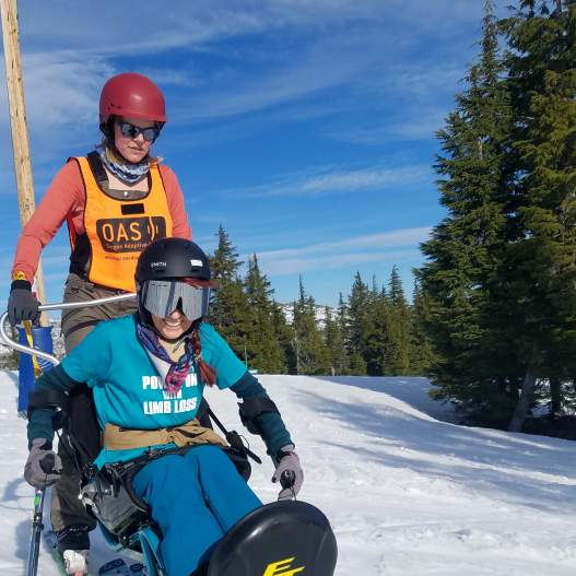 A skier uses adaptive ski equipment with a companion to ski the slopes. Snow, trees and blue sky in the backdrop.