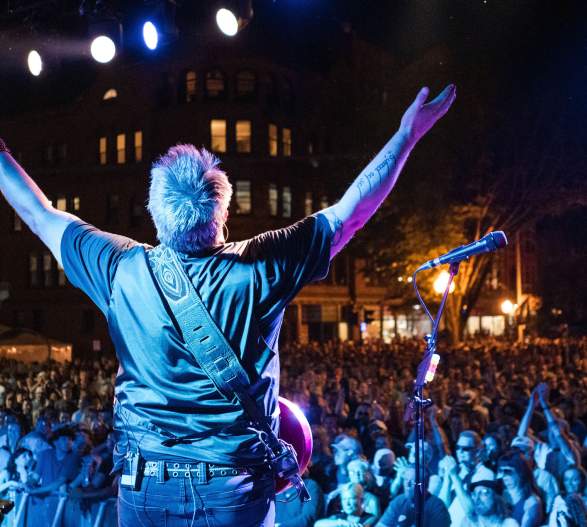 guitar player on stage with hands up in front of huge crowd at night