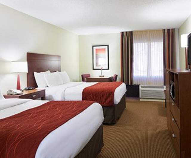 Hotel room with two beds featuring white bedding and red accent throws, wood headboards, bedside tables with lamps, a desk, dresser with TV, and a window with curtains and an in-room air-conditioning unit.