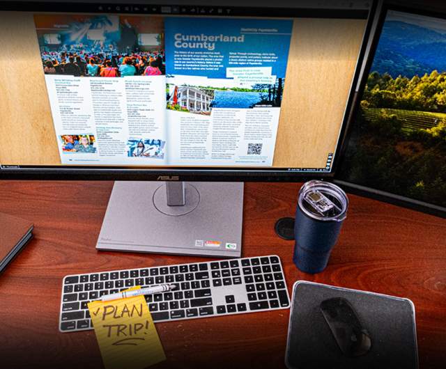 A digital destination guide is open on a computer monitor at an office desk with a keyboard, mousepad, a blue travel cup, and a yellow sticky note that says Plan Trip.