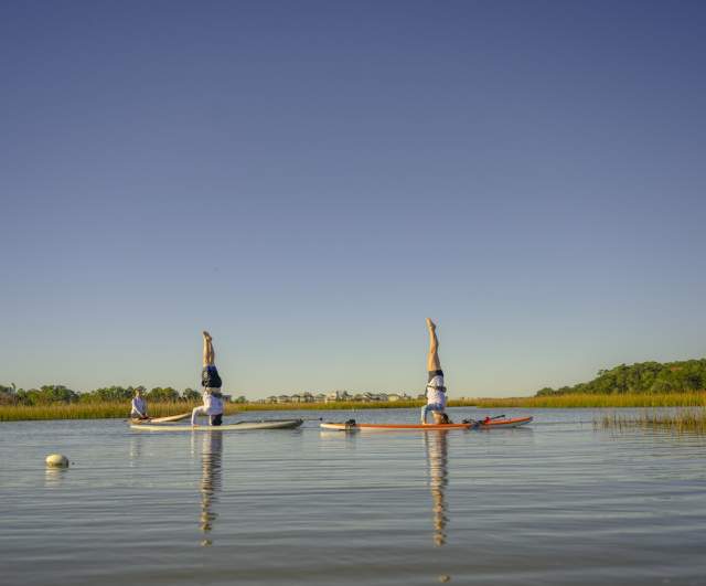 Women practicing paddleboard yoga with headstands on calm marsh waters in North Myrtle Beach, South Carolina, under a clear blue sky surrounded by peaceful natural scenery.