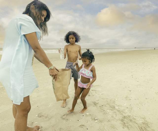 Woman and two children picking up trash on the beach, placing items into a paper bag.
