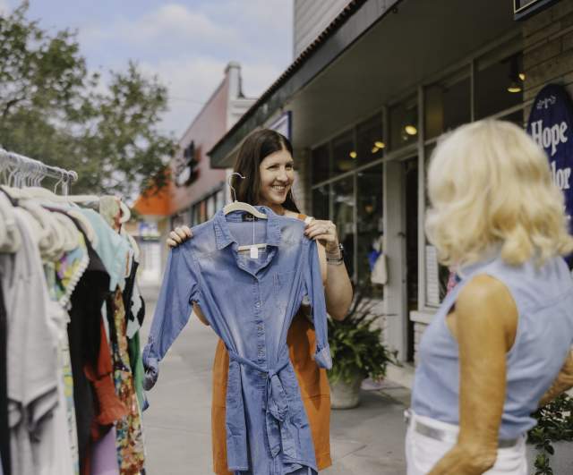A woman smiles while holding up a denim dress on a hanger outside a boutique, showing it to another woman during a sidewalk shopping trip on a sunny day.
