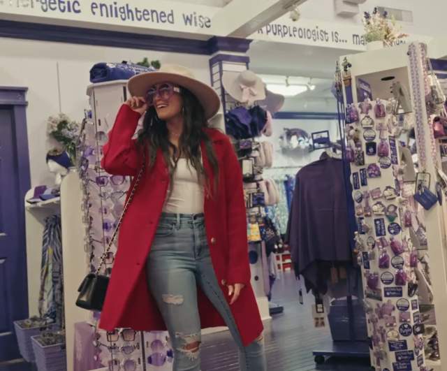 A woman wearing a red coat, wide-brimmed hat, and sunglasses browses inside a brightly lit boutique filled with purple and white décor, accessories, and gifts.