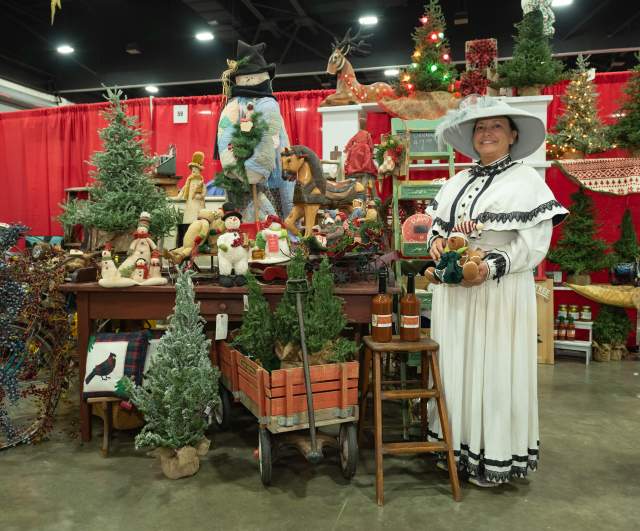 A woman wearing a white Victorian-style dress and wide-brimmed hat stands beside a festive holiday display at an indoor market. The booth features small decorated Christmas trees, wooden toys, Santa figurines, wreaths, and other holiday crafts, all set against bright red curtains.