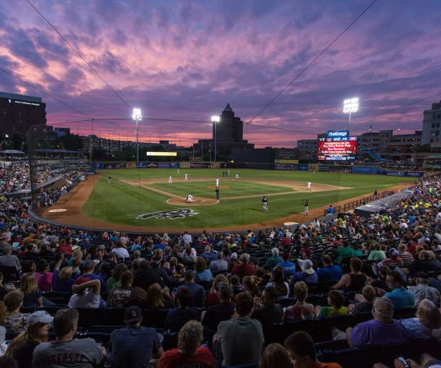 Crowded Akron Rubber Ducks baseball stadium at sunset with players on the field and a vibrant purple-pink sky overhead.