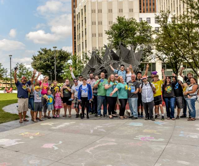 Diverse group of adults and children outdoors by modern metal sculpture and tall building in Akron area.