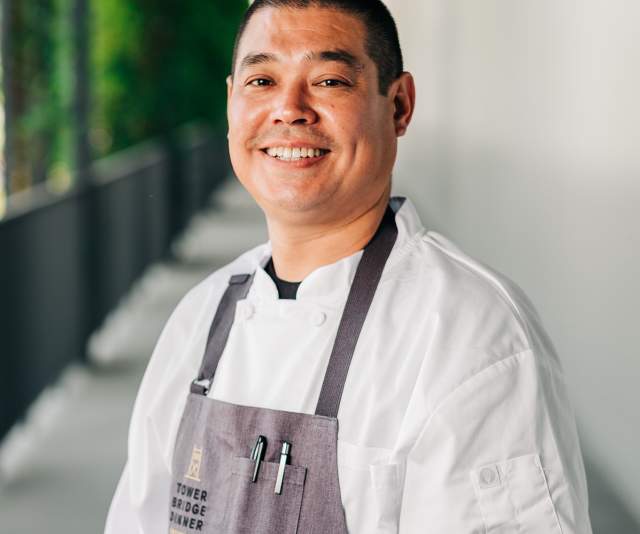 A smiling chef in a white coat and gray apron displaying the Tower Bridge Dinner logo