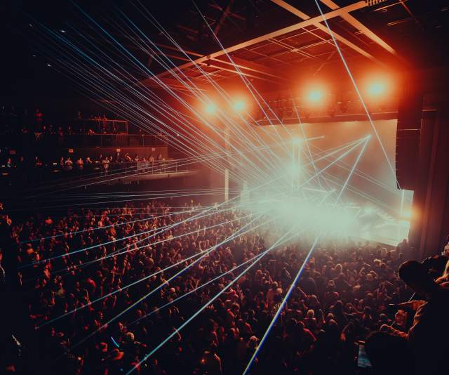 Crowd-filled concert at Channel 24 in Midtown Sacramento, with bright stage lights and blue laser beams illuminating the audience.