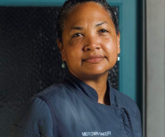 A chef in a gray overcoat smiles softly in front of a teal door