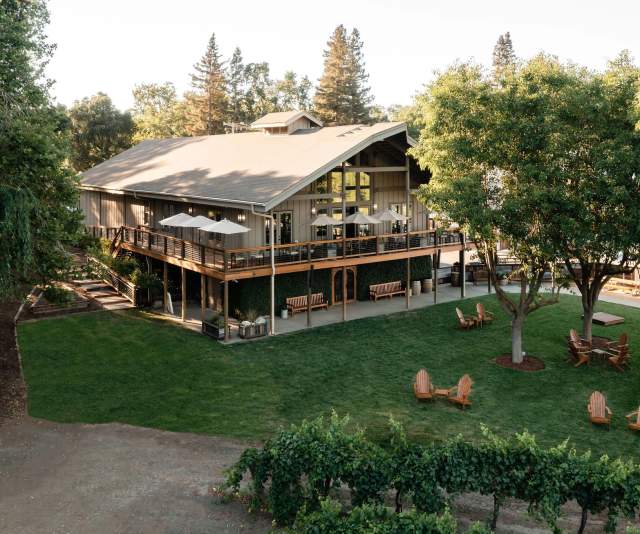 A two-story craftsman wine tasting room nestled among trees and grapevines with a manicured lawn and cozy seating areas