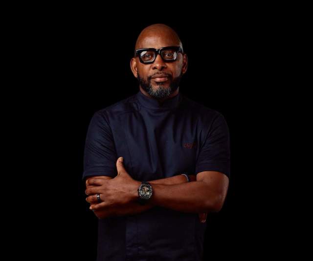 A chef in a black coat and black glasses stands confidently with arms crossed against a black background