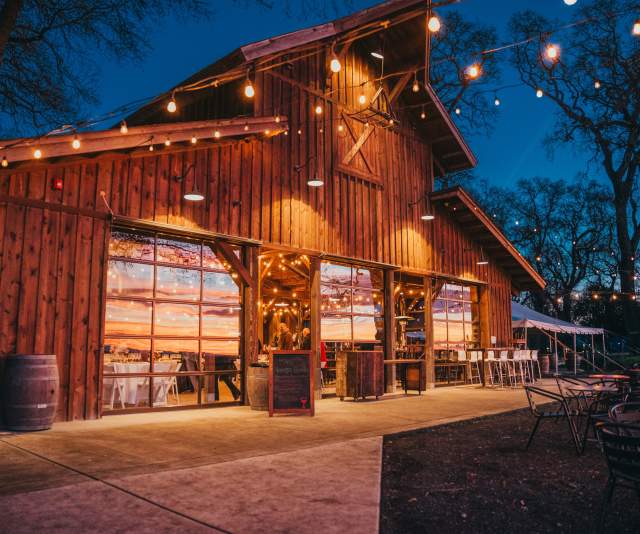 A warmly lit barn-style wine tasting room at dusk, surrounded by trees strung with market lights