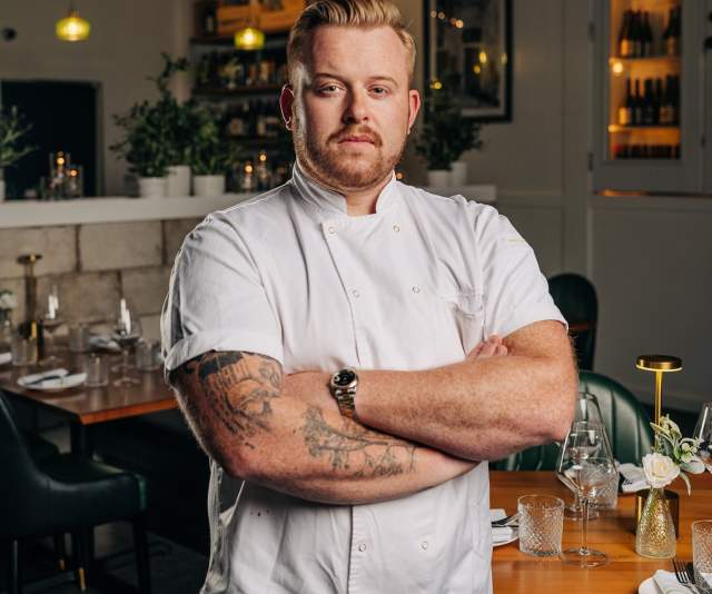 A chef in a white overcoat stands with arms crossed in a low-lit restaurant