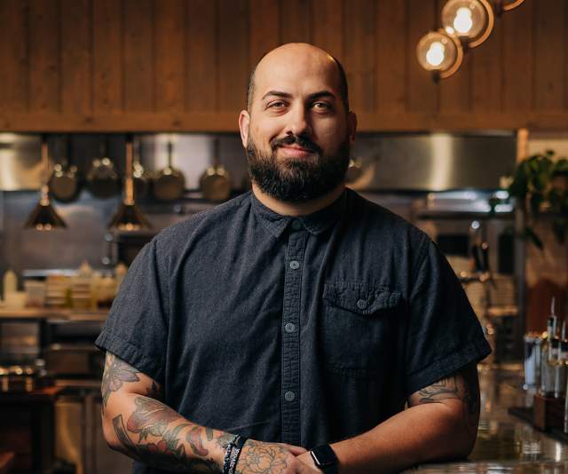 A smiling chef in a black buttoned up shirt offers a smile while leaning against the bar top inside a warmly lit restaurant