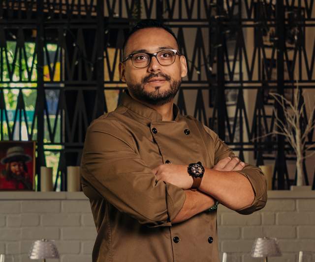 A chef in a brown overcoat stands with arms crossed in front of a black and white divider wall inside a restaurant