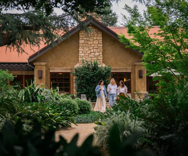 Three friends walk among a lush green garden in front of a stone tasting room while chatting and holding wine glasses