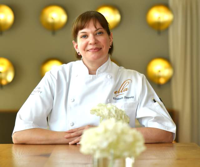 A chef in a white overcoat smiles in a warmly lit restaurant