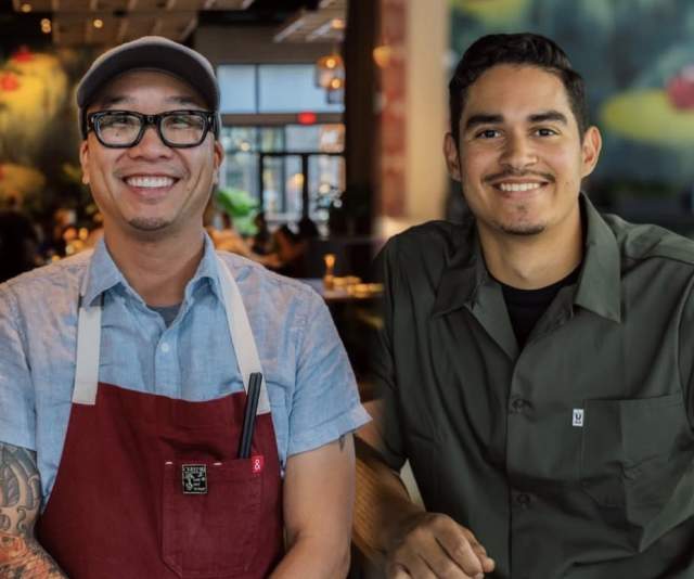A chef in a red apron and light blue collared shirt and a chef in a deep green collared shirt stand side by side in a warmly lit restaurant