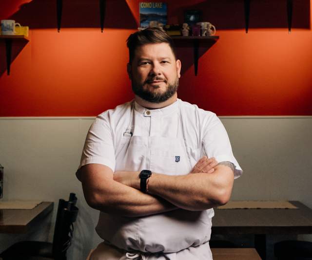 A chef in a white apron stands proudly with arms crossed while leaning against a table inside a restaurant with red and white walls