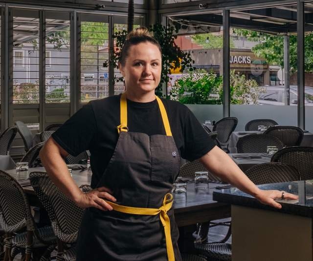 A chef in a black apron with yellow strings leans proudly against a counter inside a low-lit restaurant