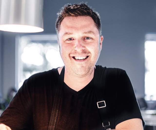 A chef in black shirt and black apron offers a smile while leaning on counter in a kitchen displaying stainless steel appliances and lighting