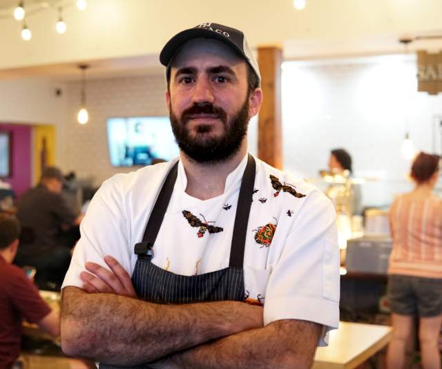 A chef in a white shirt, dark gray apron, and brown ballcap stands with arms crossed in a brightly lit restaurant
