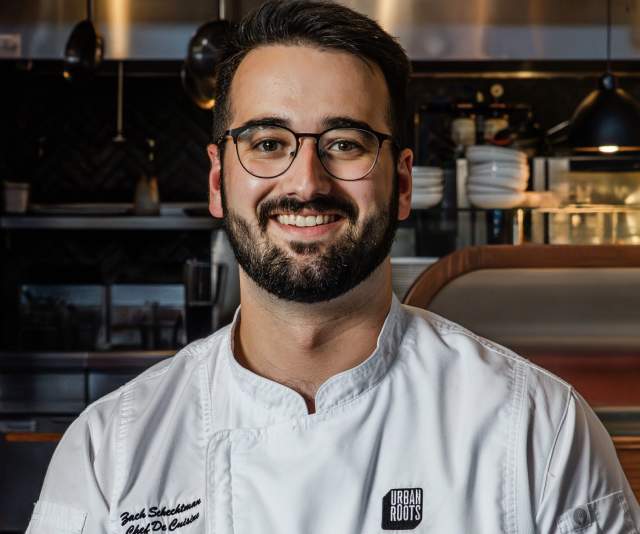 A chef in a white overcoat and glasses smiles in a stainless steel kitchen