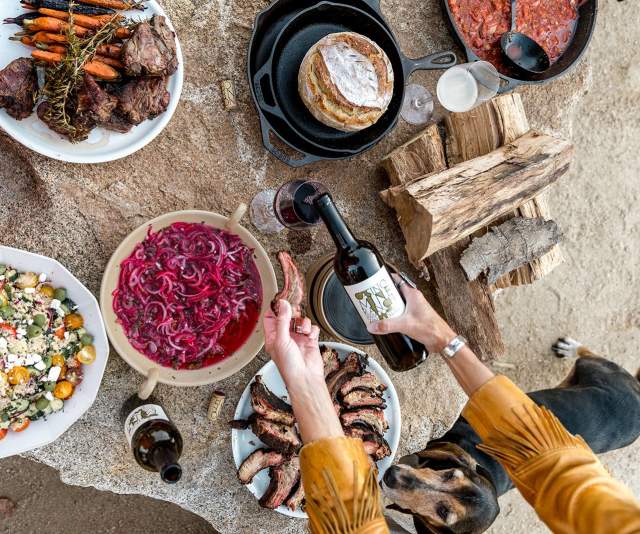 A hand pouring red wine into a glass surrounded by shareable plates of roasted meats, vegetables, and cheeses.