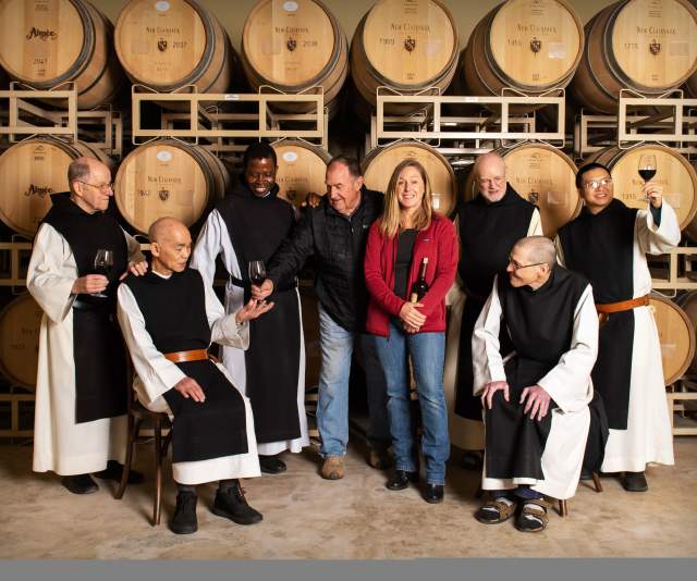 A woman in blue jeans and a red jacket and a man in blue jeans and a black jacket smile in front of a wall of wine barrels with six monks in traditional black and white belted robes