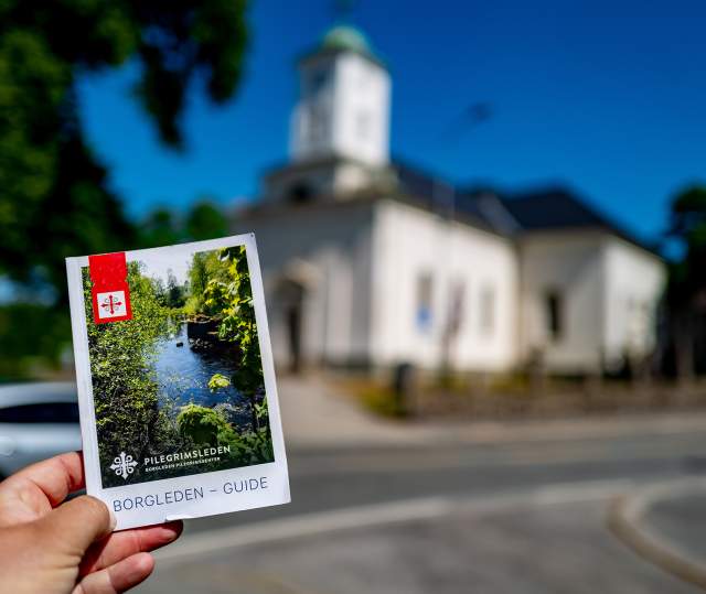 En hånd holder i en brosjyre for Borgleden foran en kirke