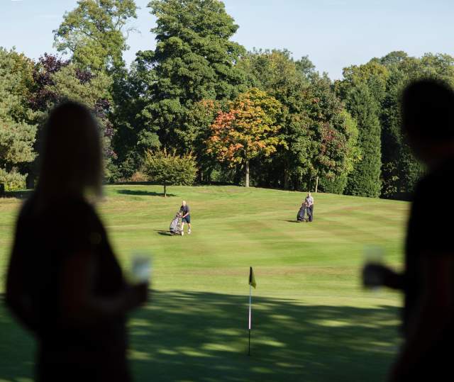 A golfing green with trees in the background and people holding drinks in the foreground
