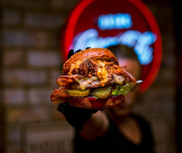 A close up of a man holding a burger up to camera in a dim lit restaurant