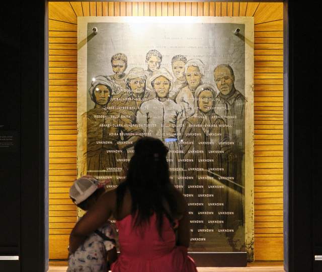 A Black woman with long hair holds her child while they look at a photo collage of formerly enslaved residents of Africatown, Alabama