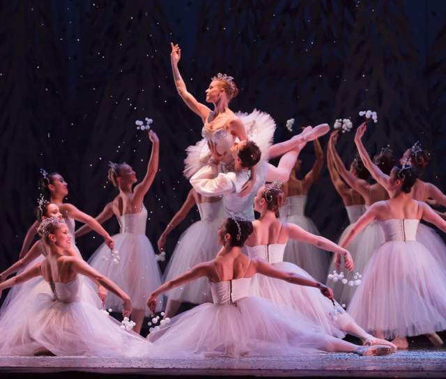 More than a dozen young girls dressed in white surround the Sugar Plum Fairy, also dressed in white in the center of the circle.