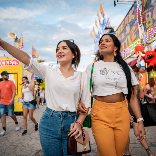 Two young women walk arm-in-arm through the Wisconsin State Fair, with one pointing excitedly ahead. Carnival rides, game booths, and crowds create a festive atmosphere in the background.
