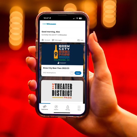 Hand holding up cellphone displaying homescreen of Milwaukee Passes in front of blurry theater background
