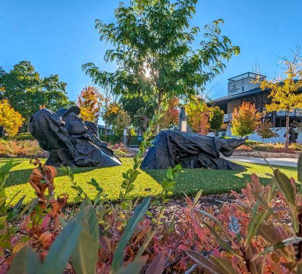 A large dark sculpture of a man lounging on a patch of green grass surrounded by a vibrant garden with a theater in the background.