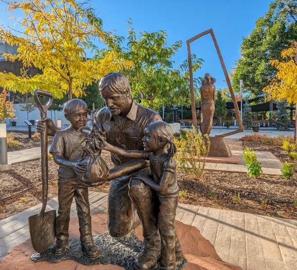 A bronze-cast sculpture of a man kneeling next to two children holding a shovel in one hand and a tree seedling in the other. The children are looking at the seedling and there are vibrant yellow fall leaves in the background.